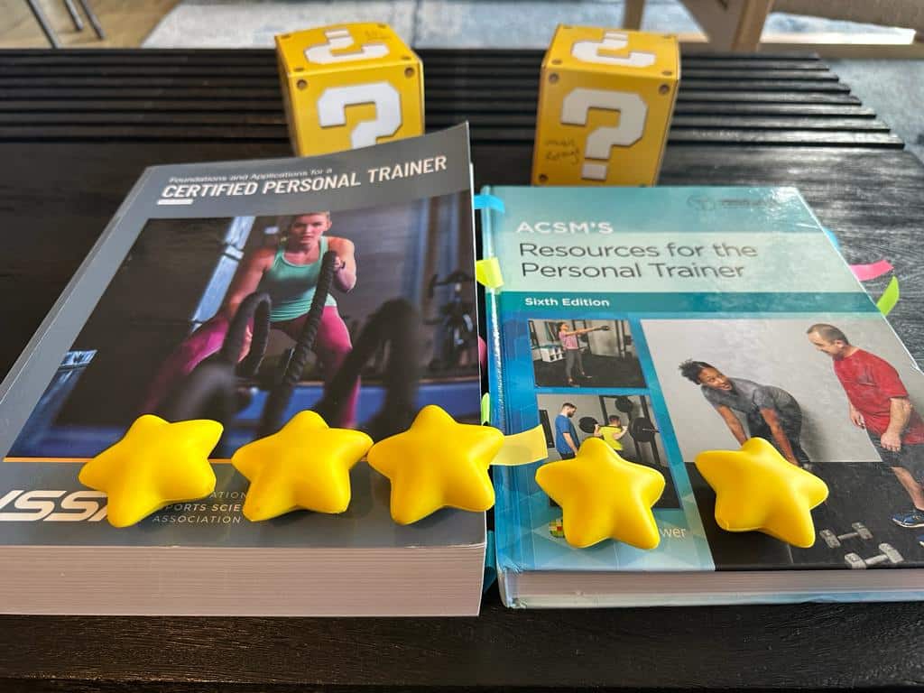 ISSA vs ACSM - Which Fitness Certification is Best? 62 ISSA and ACSM textbooks on table with gold stars and yellow question boxes