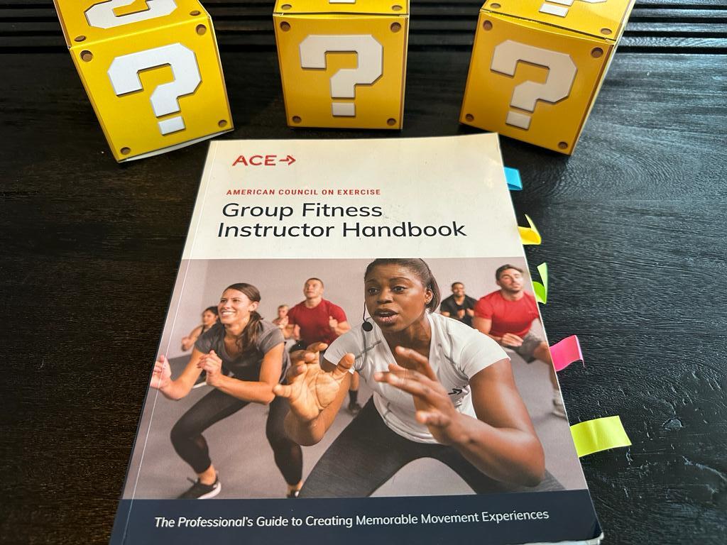 How to pick the best group fitness certification (Updated) 35 best group fitness instructor certifications - ace handbook on desk with yellow question mark boxes