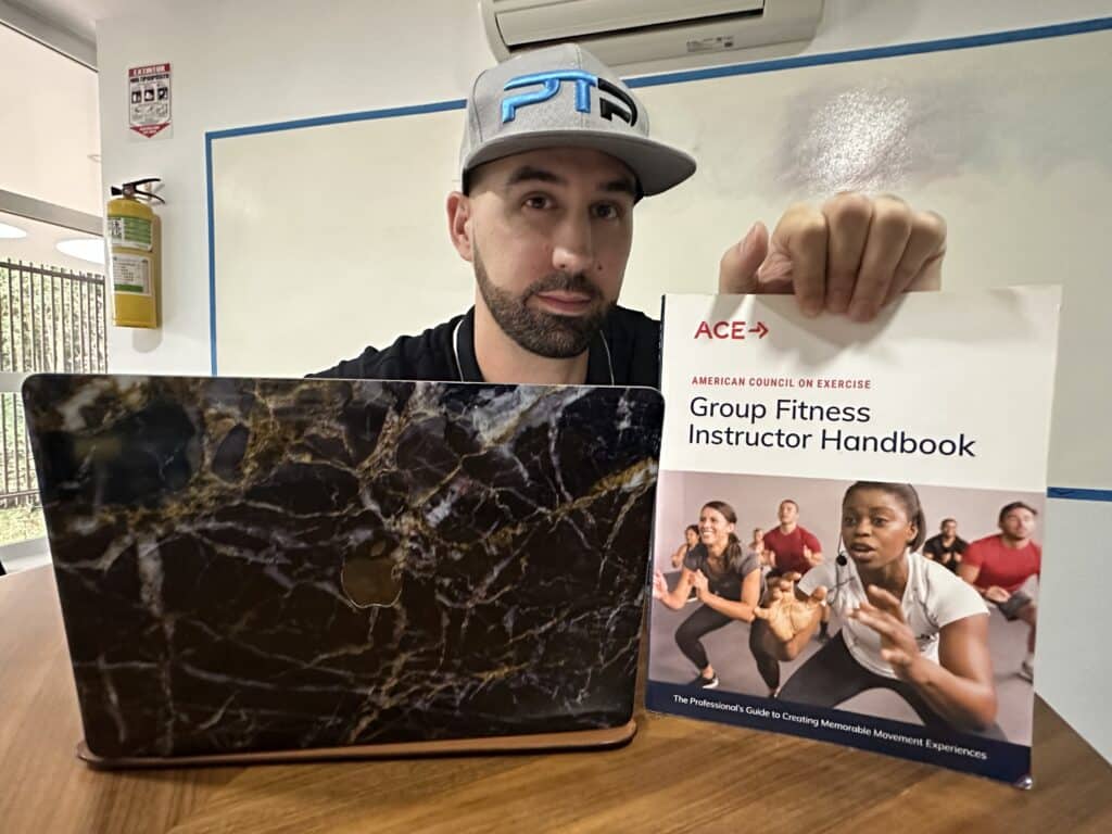 The Ultimate ACE Group Fitness Instructor Review 11 ACE Group Fitness Instructor review - Tyler read holds up ace textbook on a table next to a laptop