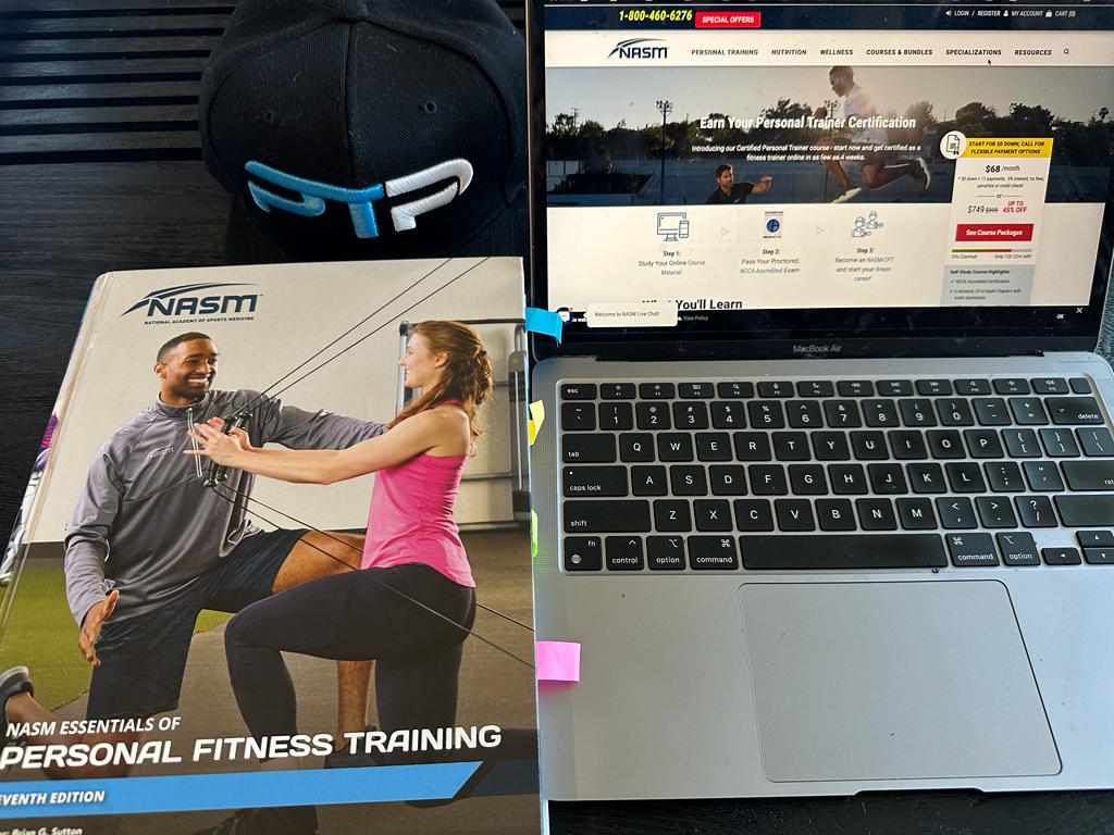 Best online personal trainer certification guide (Updated) 32 best online training certifications - NASM textbook on table next to ptpioneer hat and laptop displaying nasm website - should you pick nasm