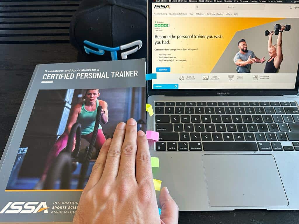 Best online personal trainer certification guide (Updated) 29 Best online personal training certification - ISSA textbook next to laptop with the issa website on the screen - ptpioneer hat and hand visible